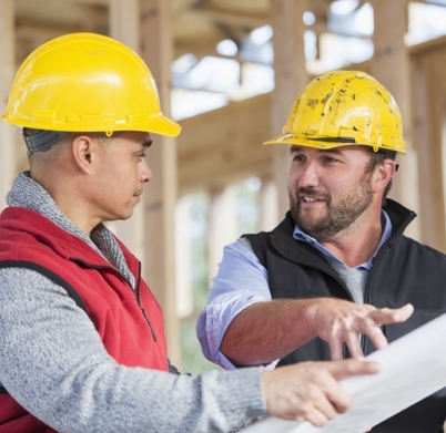 Two multi-ethnic construction workers at a job site, looking at plans. They are wearing hardhats, standing by the wood frame of a house being built. The man wearing the red vest is Hispanic. They are looking at each other, pointing and discussing the plans.